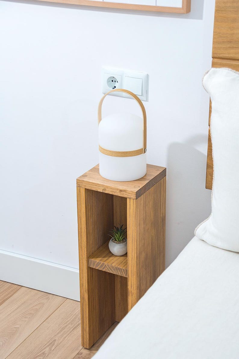 May include: A wooden bedside table with a shelf and a small plant on the shelf. A white lamp with a wooden handle sits on the top shelf.