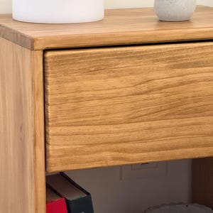 May include: A light brown wooden nightstand featuring a single drawer with a visible wood grain pattern. The top surface holds a white cylindrical container with a wooden lid and a small green succulent in a grey pot. Two books are visible below.