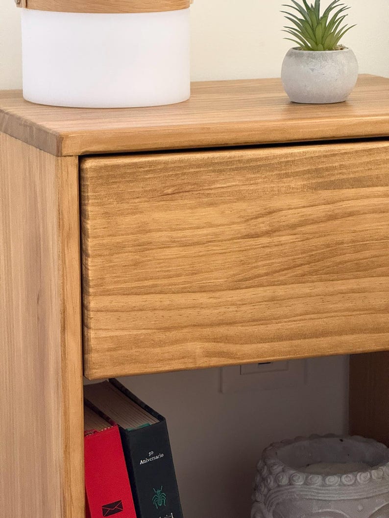 May include: A light brown wooden nightstand featuring a single drawer with a visible wood grain pattern. The top surface holds a white cylindrical container with a wooden lid and a small green succulent in a grey pot. Two books are visible below.
