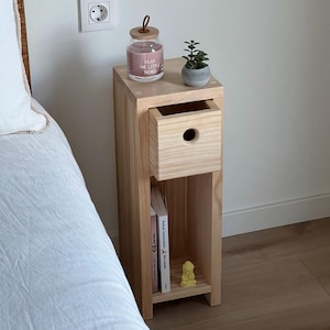 May include: A light wood bedside table with a drawer, a shelf, and a small potted succulent plant. The bedside table has a candle jar with the text "Enjoy the little things" on it.