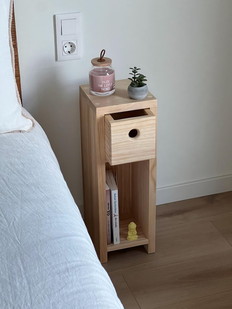 May include: A light wood bedside table with a drawer, a shelf, and a small potted succulent plant. The bedside table has a candle jar with the text "Enjoy the little things" on it.