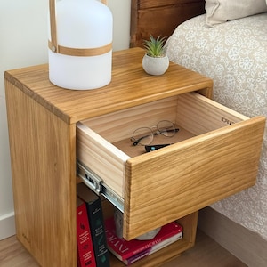 May include: A light brown wooden nightstand with a single open drawer containing a pair of round glasses. The top surface holds a white portable lamp with a wooden handle and a small potted succulent. Books are stored on the lower shelf.