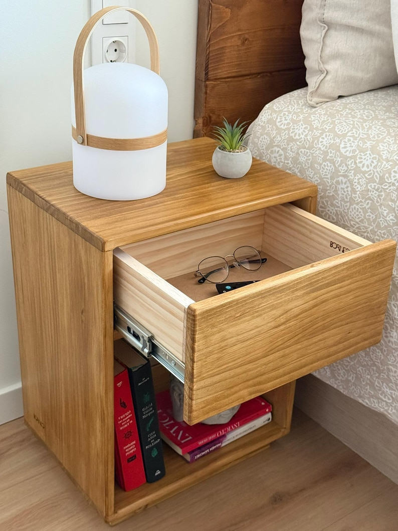 May include: A light brown wooden nightstand with a single open drawer containing a pair of round glasses. The top surface holds a white portable lamp with a wooden handle and a small potted succulent. Books are stored on the lower shelf.