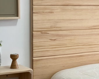 Könnte beinhalten: Ein helles Holz-Kopfteil mit horizontalen Brettern. Das Kopfteil ist an einer weißen Wand befestigt, mit einem kleinen Holzregal und einem gerahmten Kunstwerk im Hintergrund. Das Bett hat eine weiße Bettdecke.
