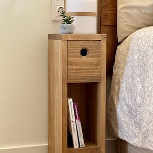 May include: A wooden bedside table with a drawer and a shelf. The table is made of light brown wood and has a simple design. There is a small potted plant on top of the table, and a lamp with a white shade is sitting on the table.