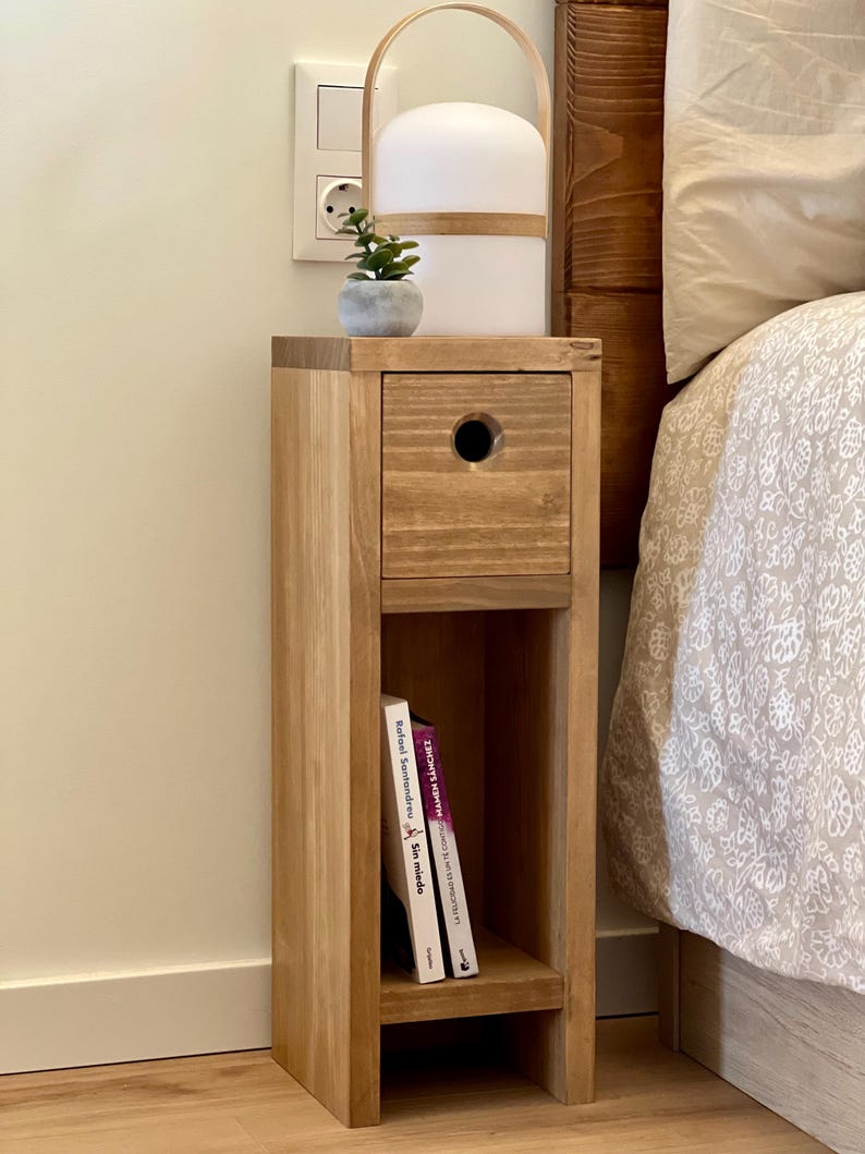 May include: A wooden bedside table with a drawer and a shelf. The table is made of light brown wood and has a simple design. There is a small potted plant on top of the table, and a lamp with a white shade is sitting on the table.