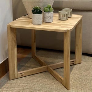 May include: A light wood end table with a square top and a cross-shaped base. The table is made of solid wood and has a natural finish. There are two small potted plants and two candle holders on the table.