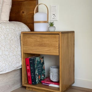 May include: A rectangular wooden nightstand with a single top drawer and an open lower shelf. The shelf contains three books with red, green, and white covers, and a gray stone-like sculpture of a face. A portable lamp and a small succulent sit on top.