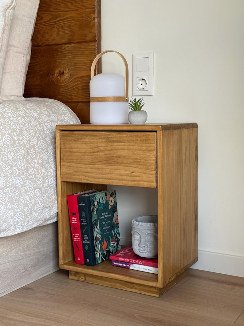 May include: A rectangular wooden nightstand with a single top drawer and an open lower shelf. The shelf contains three books with red, green, and white covers, and a gray stone-like sculpture of a face. A portable lamp and a small succulent sit on top.