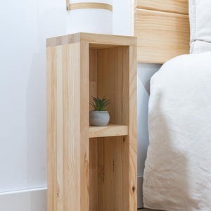 May include: A wooden bedside table with two shelves. The table is made of light wood and has a simple design. A small potted plant sits on the top shelf.