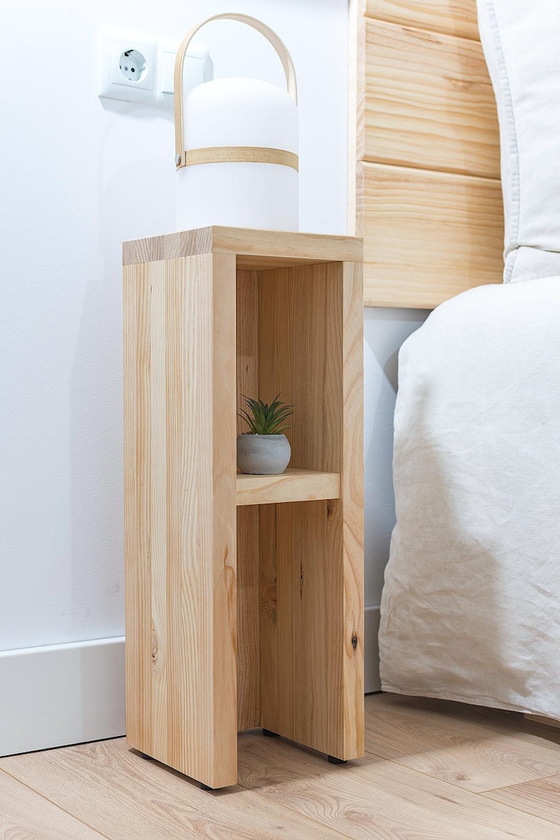 May include: A wooden bedside table with two shelves. The table is made of light wood and has a simple design. A small potted plant sits on the top shelf.