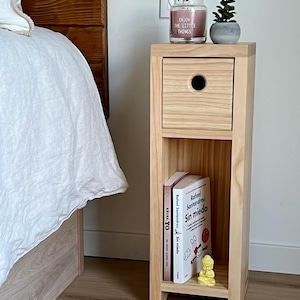 K&ouml;nnte beinhalten: Ein hoher, schmaler Nachttisch aus hellem Holz mit einer quadratischen Schublade mit kreisf&ouml;rmigem Griffloch oben und einem offenen Ablagefach darunter. Im Regal stehen zwei B&uuml;cher mit den Titeln AHORA YO und Sin miedo sowie eine kleine gelbe Figur.