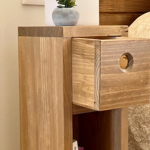 May include: A wooden bedside table with a drawer and a shelf. The drawer has a round cutout on the front. A small potted plant sits on the top shelf.