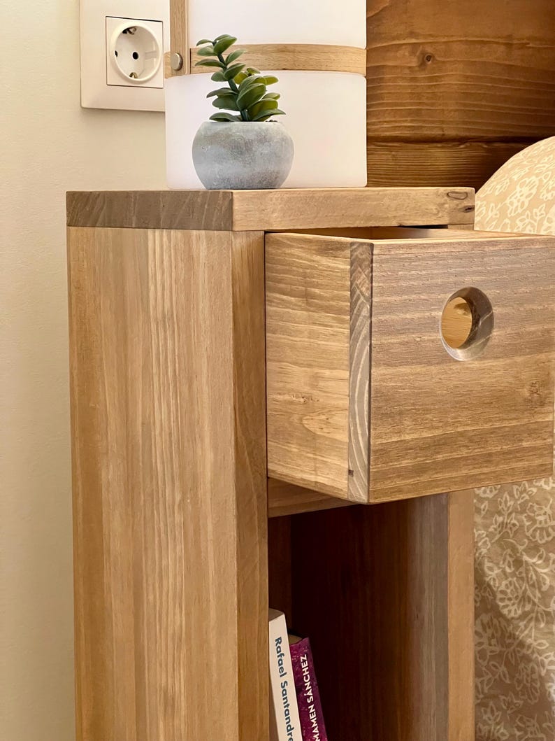 May include: A wooden bedside table with a drawer and a shelf. The drawer has a round cutout on the front. A small potted plant sits on the top shelf.