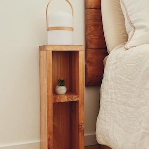 K&ouml;nnte beinhalten: Ein hoher, schmaler Nachttisch aus Holz mit hellbrauner Oberfl&auml;che, der eine flache Oberseite und ein offenes Regal besitzt. Eine wei&szlig;e, kuppelf&ouml;rmige, tragbare Lampe mit hellem Griff steht oben darauf, und eine kleine gr&uuml;ne Sukkulente in einem Topf steht im Regal.