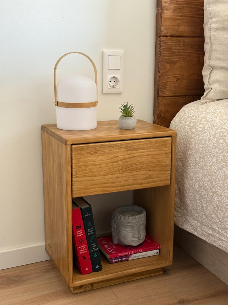 May include: A light brown wooden nightstand with a single drawer and an open lower shelf. On top sits a white portable lamp with a light wood handle and a small succulent in a gray pot. The lower shelf holds two books and a gray Buddha head statue.