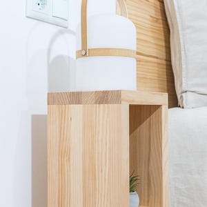 May include: A wooden bedside table with two shelves. The top shelf has a white lamp with a wooden handle. The bottom shelf has a small potted plant.
