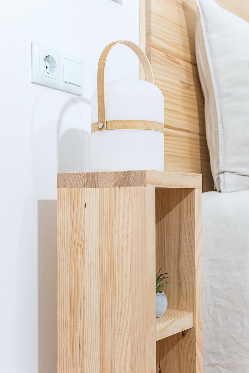 May include: A wooden bedside table with two shelves. The top shelf has a white lamp with a wooden handle. The bottom shelf has a small potted plant.