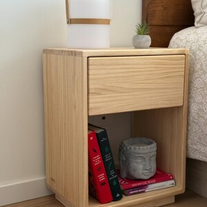 May include: A light-colored wooden nightstand with a single drawer and an open storage shelf below. The shelf contains three books with red, green, and pink spines, and a gray stone-like Buddha head sculpture. A portable white lamp sits on top.