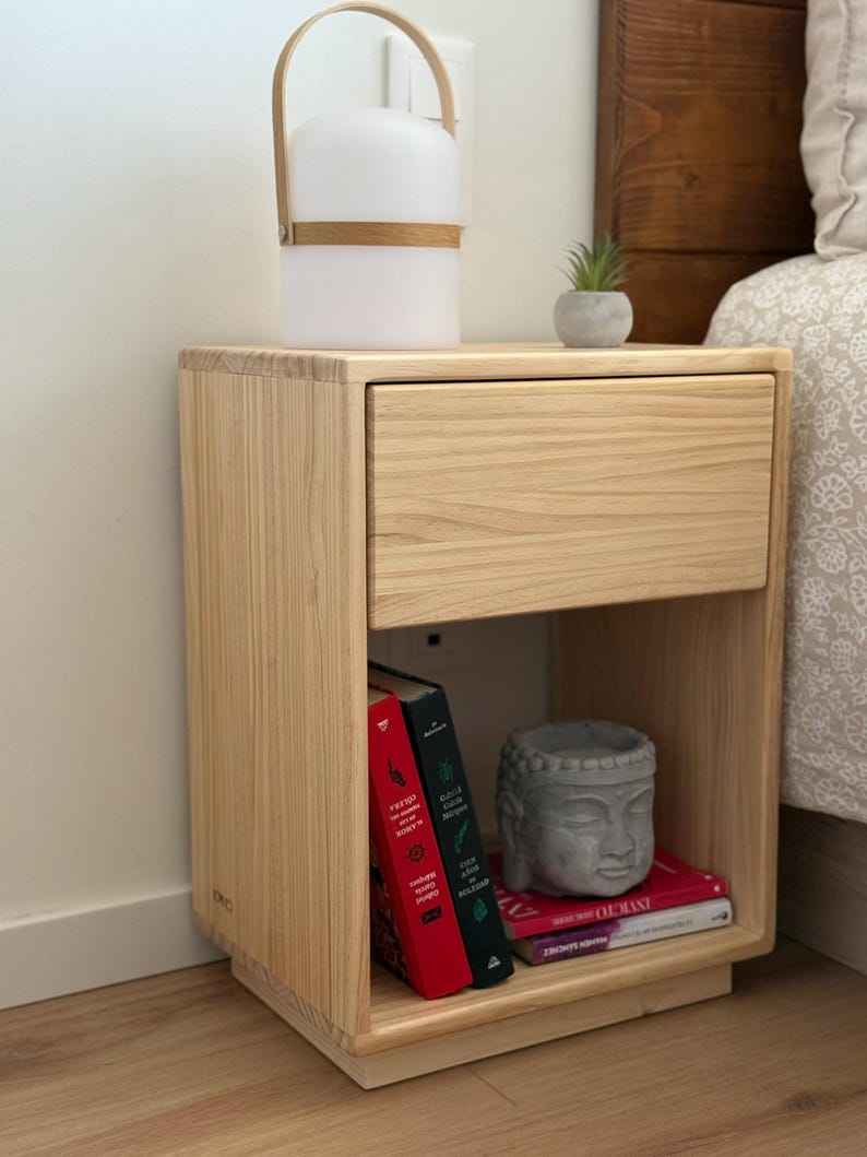 May include: A light-colored wooden nightstand with a single drawer and an open storage shelf below. The shelf contains three books with red, green, and pink spines, and a gray stone-like Buddha head sculpture. A portable white lamp sits on top.
