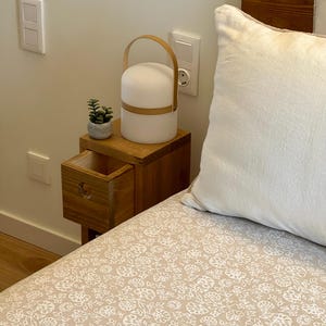 May include: A wooden bedside table with a drawer, a small potted plant, and a white lamp with a wooden handle. The table is next to a bed with a white pillow and a beige floral patterned bedspread.