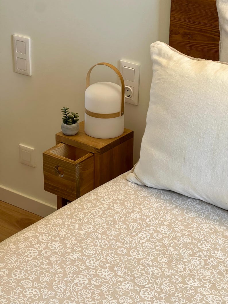 May include: A wooden bedside table with a drawer, a small potted plant, and a white lamp with a wooden handle. The table is next to a bed with a white pillow and a beige floral patterned bedspread.