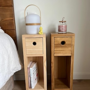 May include: Two wooden bedside tables with a drawer and a shelf. The bedside table on the left has a white lamp with a wooden handle and a small yellow figurine on top. The bedside table on the right has a glass jar candle with the text "Enjoy the little things".