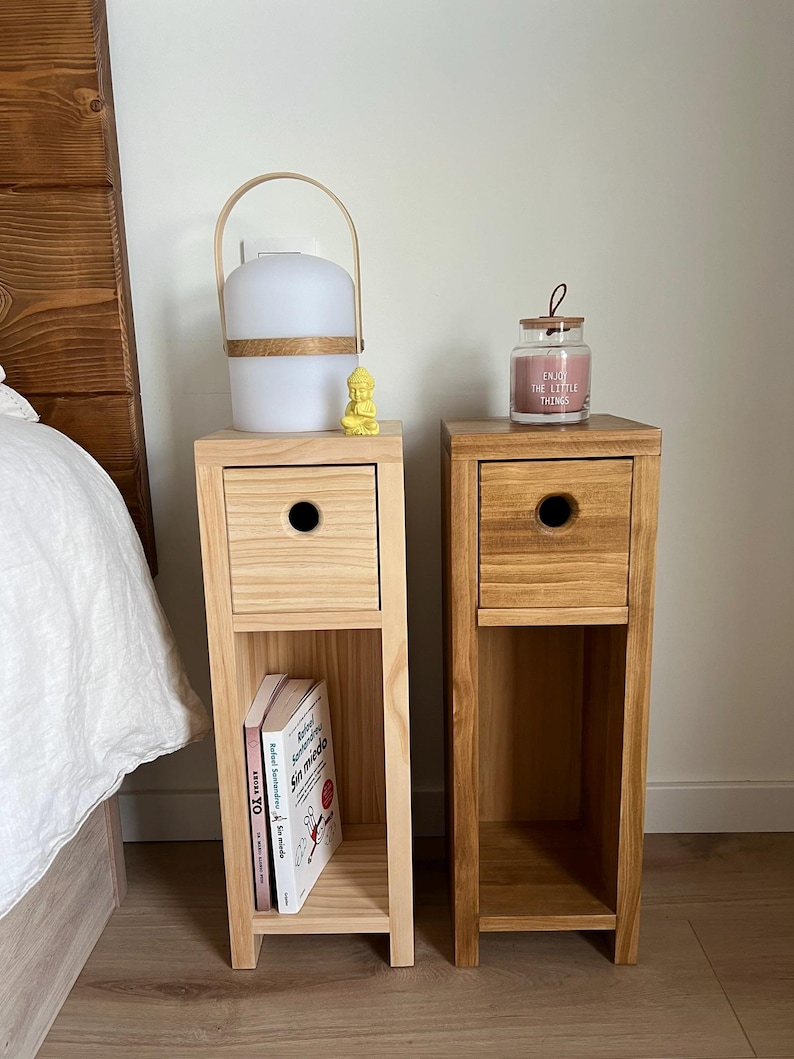 May include: Two wooden bedside tables with a drawer and a shelf. The bedside table on the left has a white lamp with a wooden handle and a small yellow figurine on top. The bedside table on the right has a glass jar candle with the text "Enjoy the little things".