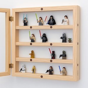 Puede incluir: Una vitrina de madera con puerta de cristal, que muestra varias minifiguras de Lego de Star Wars. La vitrina tiene varios estantes y está montada en una pared blanca. Las minifiguras incluyen personajes como Darth Vader y Yoda.