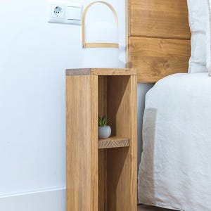 May include: A wooden bedside table with two shelves. The table is stained a light brown color and has a simple design. A small potted plant sits on the top shelf.