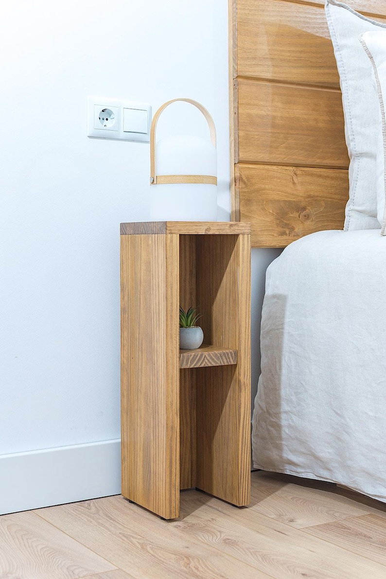 May include: A wooden bedside table with two shelves. The table is stained a light brown color and has a simple design. A small potted plant sits on the top shelf.