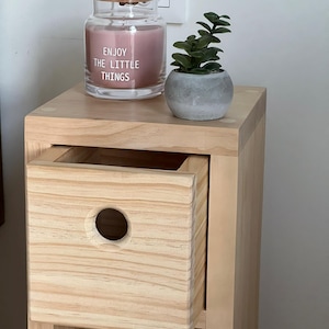 May include: A light wood bedside table with a drawer and a shelf. The drawer has a round cutout. A pink candle in a glass jar with a wooden lid and the text "Enjoy the little things" is on the top shelf. A small potted plant is on the top shelf.