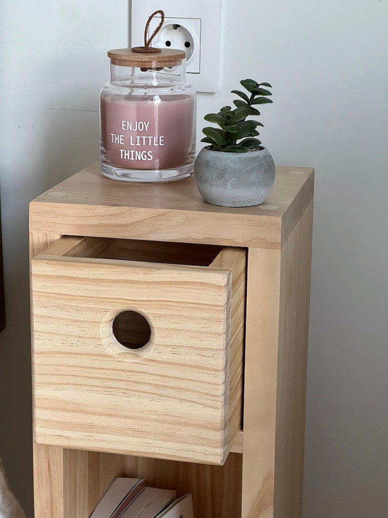 May include: A light wood bedside table with a drawer and a shelf. The drawer has a round cutout. A pink candle in a glass jar with a wooden lid and the text "Enjoy the little things" is on the top shelf. A small potted plant is on the top shelf.
