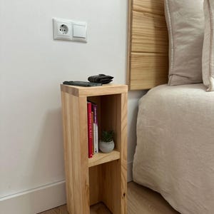 May include: A light wood bedside table with two shelves. The top shelf has a small potted plant and a wallet. The bottom shelf has a stack of books. The table is next to a bed with a white headboard and a white bedspread.