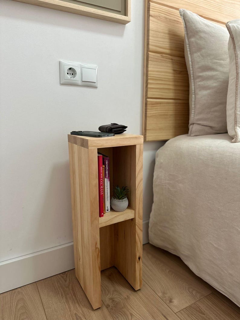 May include: A light wood bedside table with two shelves. The top shelf has a small potted plant and a wallet. The bottom shelf has a stack of books. The table is next to a bed with a white headboard and a white bedspread.