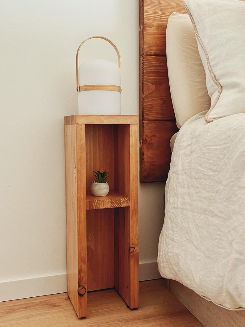 May include: A wooden bedside table with two shelves. The top shelf has a white lamp with a wooden handle. The bottom shelf has a small potted plant. The table is next to a bed with white linens.