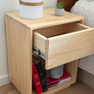May include: A light-colored wooden nightstand featuring a single pull-out drawer with metal glides and an open storage shelf below. The shelf contains two upright books with red and black spines and two stacked books with red covers, plus a gray stone head.