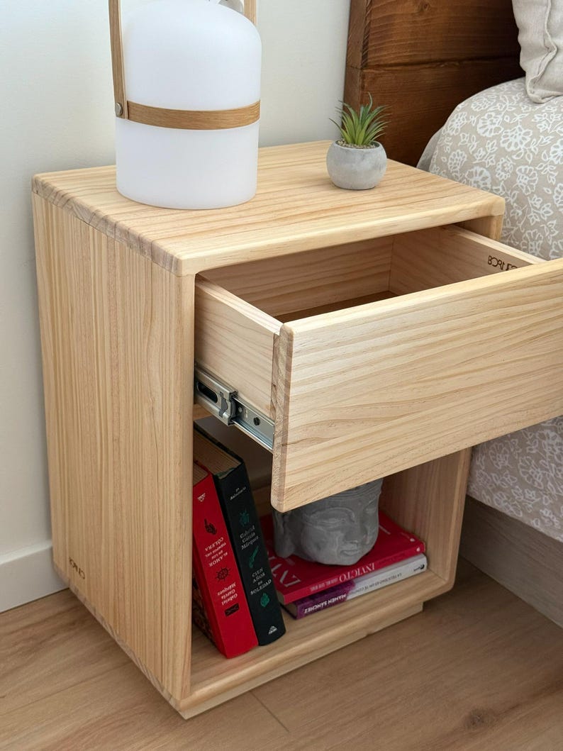 May include: A light-colored wooden nightstand featuring a single pull-out drawer with metal glides and an open storage shelf below. The shelf contains two upright books with red and black spines and two stacked books with red covers, plus a gray stone head.