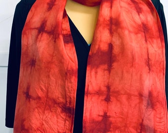 May include: Red silk scarf with a tie-dye pattern. The scarf is folded and draped over a black shirt. The scarf has a tag that says "Mien" and "Hand dyed silk scarf" with a price of $25.