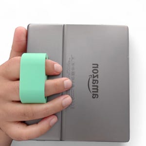 Puede incluir: Un lector electrónico gris con el logotipo de Amazon en la parte posterior. Un bucle para el dedo de color verde claro está unido a la parte posterior del lector electrónico.