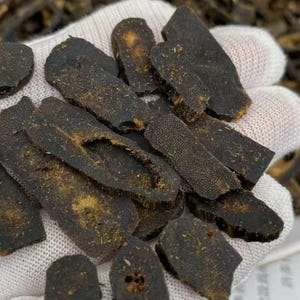 May include: Close-up of dark brown, sliced pieces of a dried root, possibly for herbal remedies. The pieces vary in shape and size, with visible textures and a light dusting of what appears to be dried herbs. The background is a white glove.