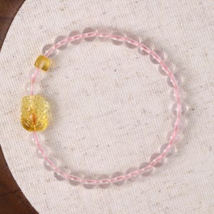 Könnte beinhalten: Ein zartes Armband mit durchscheinenden rosa Quarzperlen, auf einem rosa elastischen Band aufgereiht. Das Armband enthält einen geschnitzten gelben Edelsteinanhänger und eine gelbe rechteckige Perle, was ein einzigartiges und elegantes Accessoire ergibt.