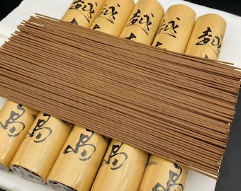 May include: A collection of brown incense sticks arranged over several rolled bundles. The bundles are wrapped in tan paper with black Japanese characters. The incense sticks are a light brown color and appear to be made of wood.