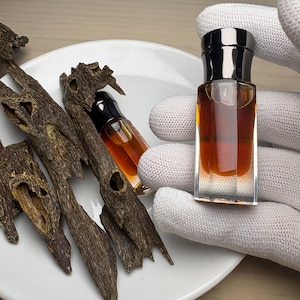 May include: A small, clear glass bottle filled with amber-colored liquid, held by a gloved hand. The bottle has a black cap. Beside it are pieces of dark brown wood on a white plate, suggesting a fragrance or essential oil product.