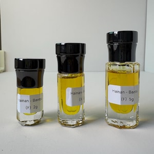 May include: Three small glass bottles with black lids, filled with a yellow liquid. Each bottle has a white label with the text "Hainan - Bartou (F)" and weights of 2g and 5g. The bottles are arranged in a row.