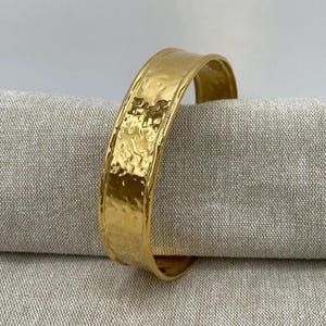 Könnte beinhalten: Ein goldfarbenes Armreif-Armband mit gehammerter Textur. Das Armband hat ein breites Band mit leicht gebogener Form. Die Oberfläche des Armbands hat ein strukturiertes, gehämmertes Aussehen und reflektiert das Licht. Das Armband wird auf einem neutralfarbenen Stoff präsentiert.