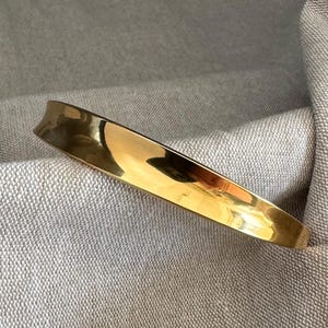 Könnte beinhalten: Ein poliertes, goldfarbenes Metallarmband mit geschwungenem Design. Das Armband hat eine glatte, reflektierende Oberfläche und liegt vor einem neutralfarbenen Stoffhintergrund. Das Design des Armbands ist schlicht und elegant.
