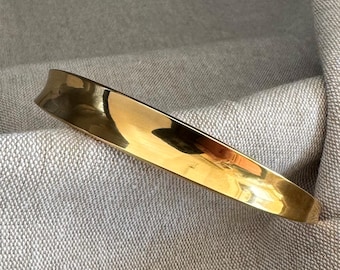 May include: A polished gold-tone metal bracelet with a curved design. The bracelet has a smooth, reflective surface and is set against a neutral-toned fabric background. The bracelet's design is simple and elegant.
