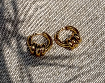 May include: A pair of gold-colored hoop earrings. Each earring features a series of three stacked, rounded segments. The earrings are displayed on a textured, neutral-toned surface, with shadows adding depth.