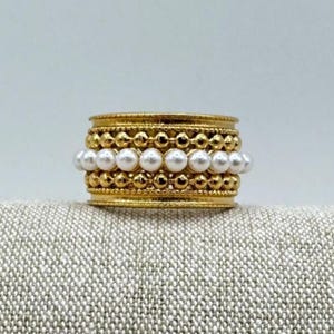 Könnte beinhalten: Ein goldfarbener Ring mit Banddesign. Der Ring hat drei Reihen von Verzierungen: kleine goldene Perlen, weiße Perlen und weitere kleine goldene Perlen. Der Ring ist vor einem neutralen Hintergrund platziert.